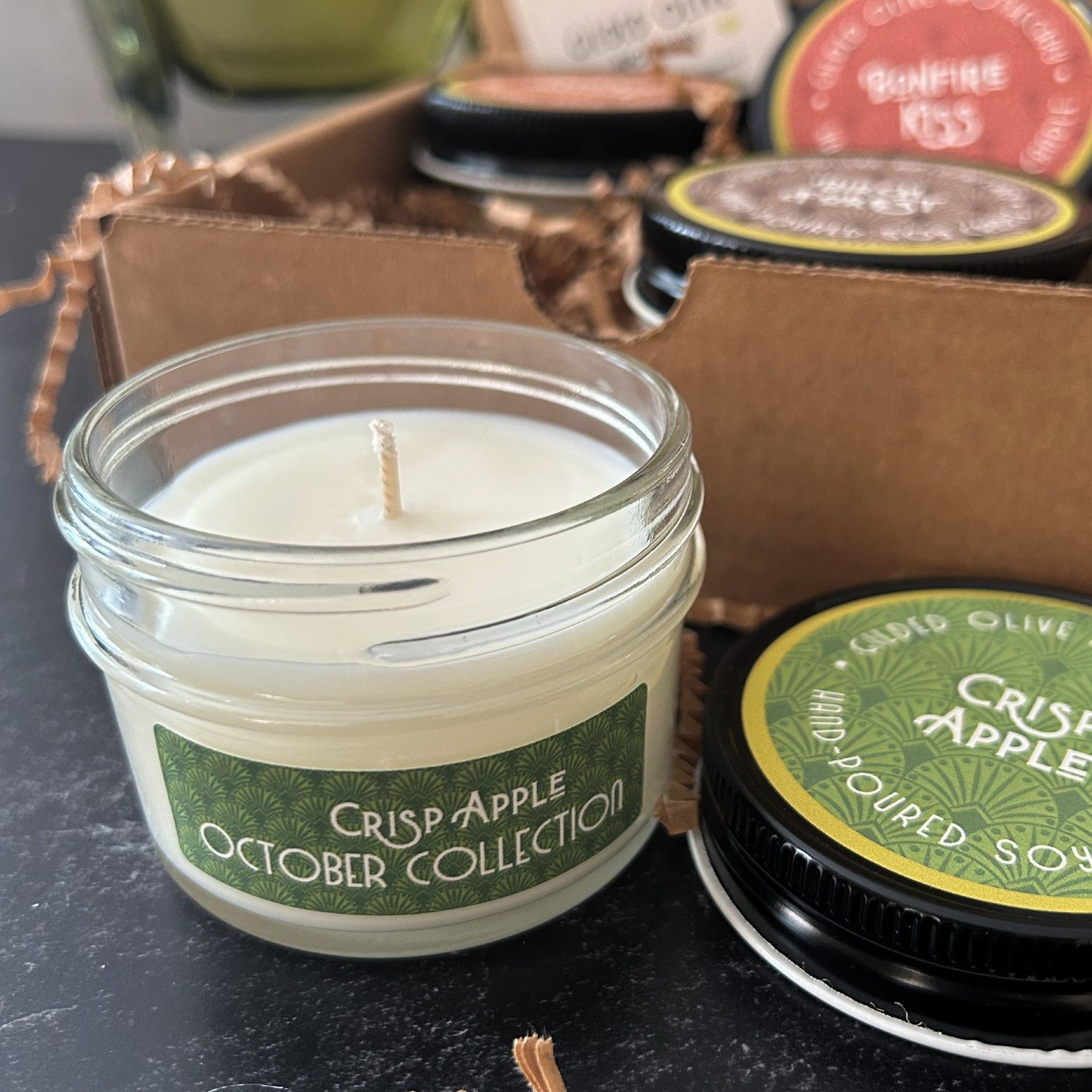 Candle in a jar labeled 'Crisp Apple October Collection' with a kraft gift box in the background.