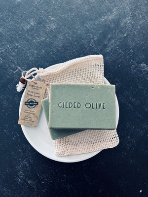 A bar of handmade soap resting on a white plate with a beige cotton soap saver bag and a kraft paper tag. 