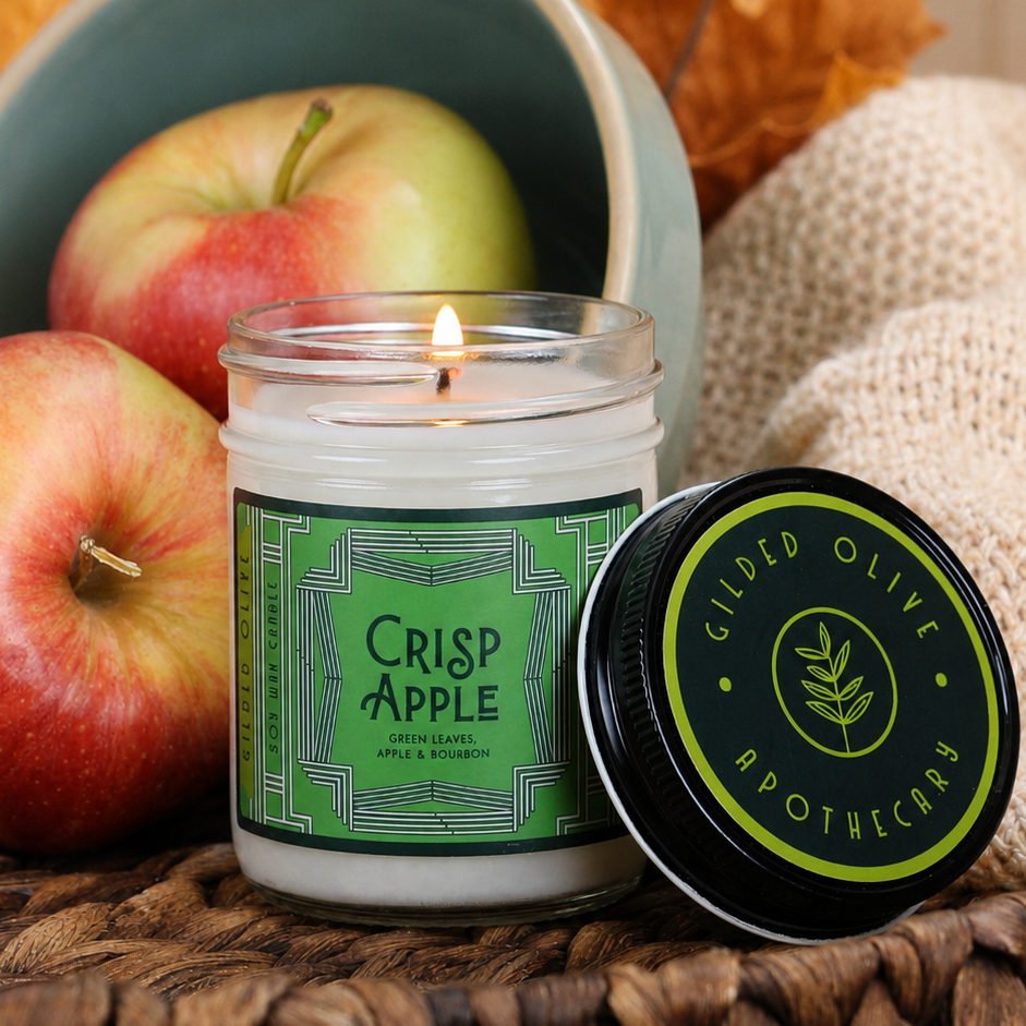 A soy wax candle called "Crisp Apple" sits on a woven brown surface. Apples and burlap fabric create a styled seasonal scene.