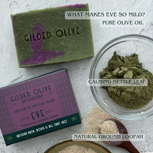 A styled flat-lay of Gilded Olive Apothecary 'Eve' loofah soap, showing the green and purple wrapped soap bar, its purple patterned box, a glass bowl of green nettle leaf powder, and natural ground loofah on a textured white background with descriptive text overlays.