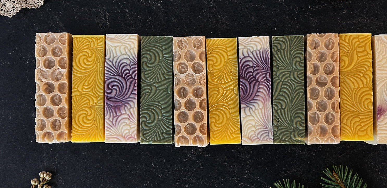 Handmade soap bars arranged on a dark textured surface. The soaps feature three repeating patterns: a honeycomb texture, a yellow swirl pattern, and a cream swirl pattern with purple accents.