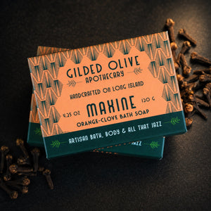 A handmade boxed bath soap product labeled ''Maxine Orange-Clove Bath Soap,' photographed on a dark surface with scattered cloves around it. 