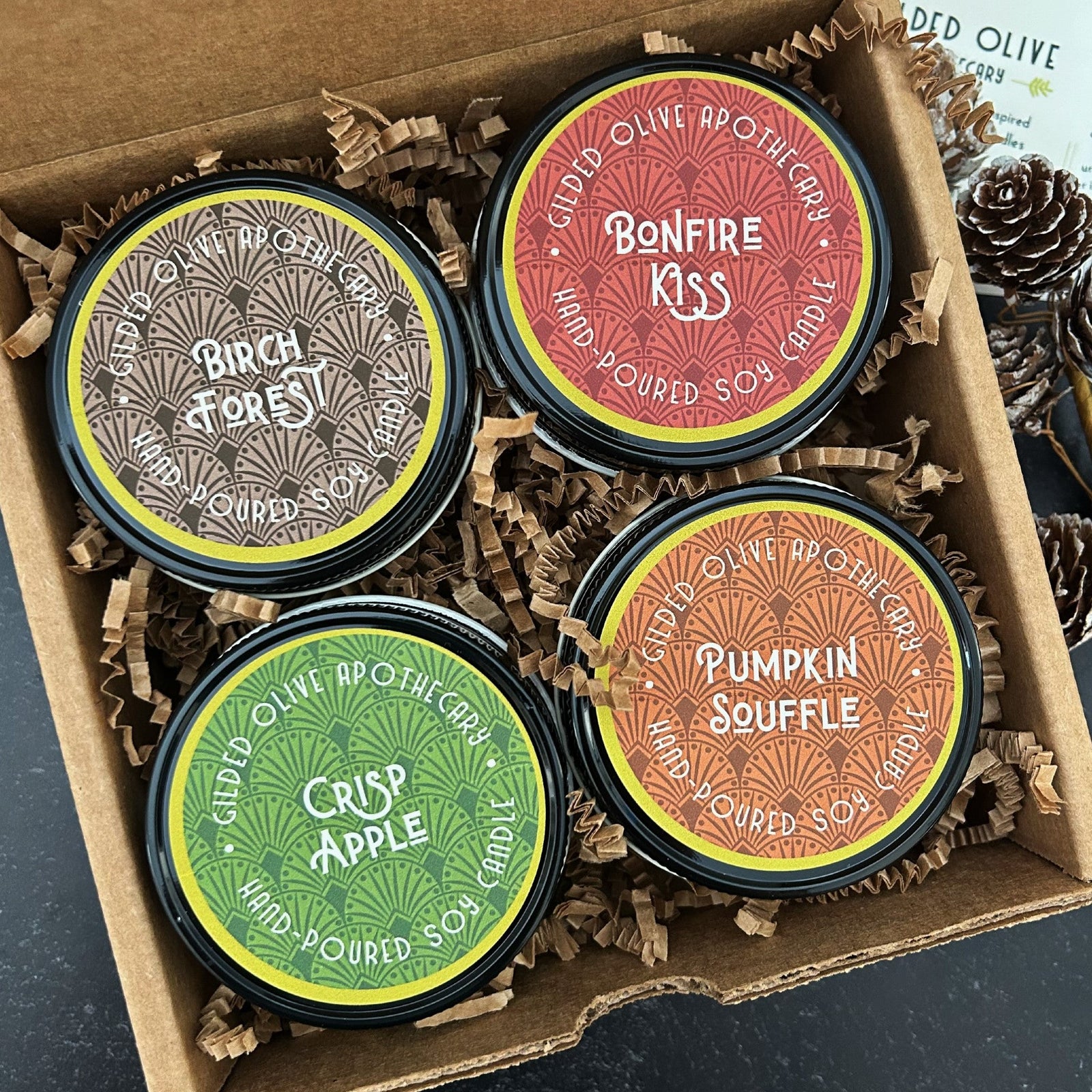 October Collection 4 oz candles in a box with paper shreds, featuring 'Birch Forest', 'Bonfire Kiss', 'Pumpkin Souffle', and 'Crisp Apple' fall scents.