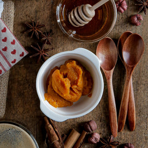 A white ceramic bowl filled with orange pumpkin puree, styled on a rustic wooden surface with wooden spoons, a glass of honey, star anise, cinnamon sticks, and a heart-patterned cloth.