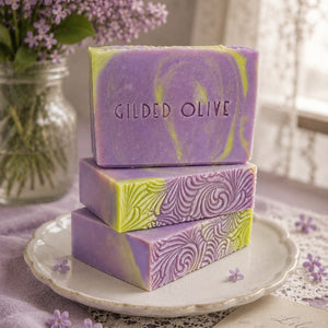 Three bars of handmade soap stacked on a vintage plate with lilac flowers in the background.
