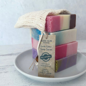 Stack of handmade soap bars with a cotton soap saver bag on a white plate against a light gray background